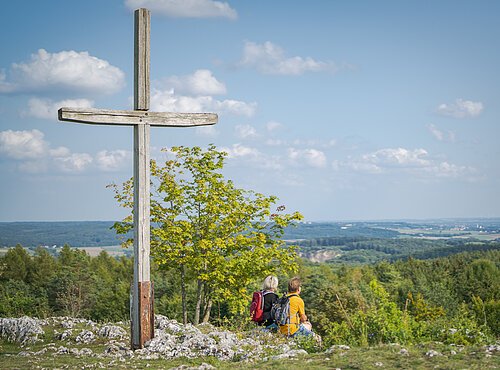 Zwei Wanderer sitzen neben dem Gipfelkreuz auf dem Bockberg und genießen die Aussicht
