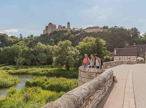 Drei Wanderer stehen auf der steinernen Brücke und blicken auf die Wörnitz. Links oben thront über dem Wald die Harburg.
