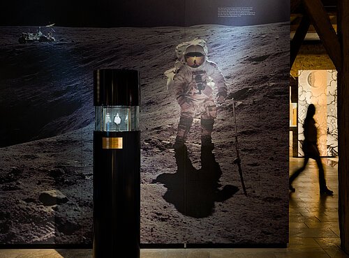 Foto von einem Astronaut, der auf dem Mond unterwegs ist. Im Vordergrund ist in der Vitrine ein Stein zu sehen. Vermutlich ein Mondstein.