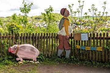 Hans im Glück, dargestellt durch eine Figur mit Gans unter dem Arm und ein Schwein aus Holz. Im Hintergrund sieht man die Stadt Harburg