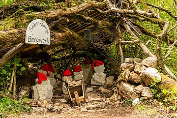 Ein Bergwerkstollen wird durch einen Gang, gebildet durch Äste dargestellt. Darin stehen Zwergenfiguren aus Stein mit roten Mützen. Rechts im Stollen sieht man eine Spinne im Netz.
