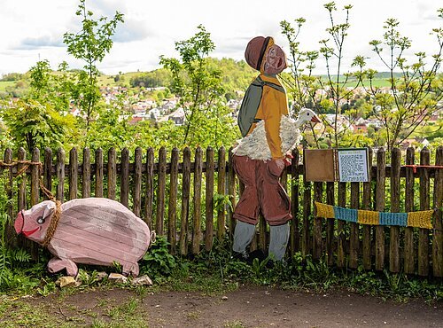 Hans im Glück, dargestellt durch eine Figur mit Gans unter dem Arm und ein Schwein aus Holz. Im Hintergrund sieht man die Stadt Harburg