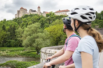 Zwei Personen mit Fahrrädern und Helmen stehen auf einer Brücke und blicken auf eine Burg auf einem Hügel.