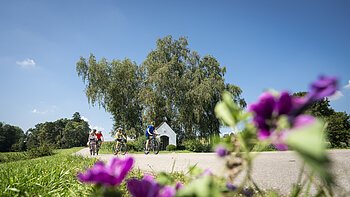 Drei Personen fahren auf Fahrrädern auf einem Weg, im Hintergrund Bäume und eine kleine Kapelle. Im Vordergrund unscharfe Blumen.