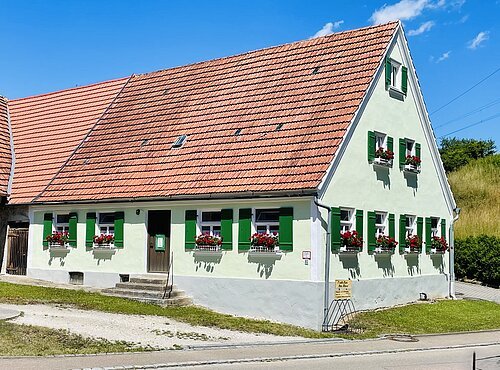 Weißes Haus mit grün lackierten Fensterläden und roten Blumen in Blumenkästen unter einem roten Ziegeldach bei blauem Himmel.