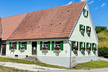 Weißes Haus mit grün lackierten Fensterläden und roten Blumen in Blumenkästen unter einem roten Ziegeldach bei blauem Himmel.