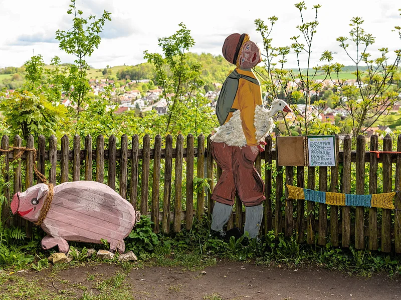 Hans im Glück, dargestellt durch eine Figur mit Gans unter dem Arm und ein Schwein aus Holz. Im Hintergrund sieht man die Stadt Harburg