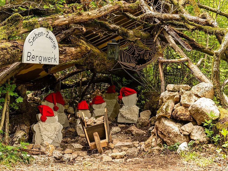 Ein Bergwerkstollen wird durch einen Gang, gebildet durch Äste dargestellt. Darin stehen Zwergenfiguren aus Stein mit roten Mützen. Rechts im Stollen sieht man eine Spinne im Netz.
