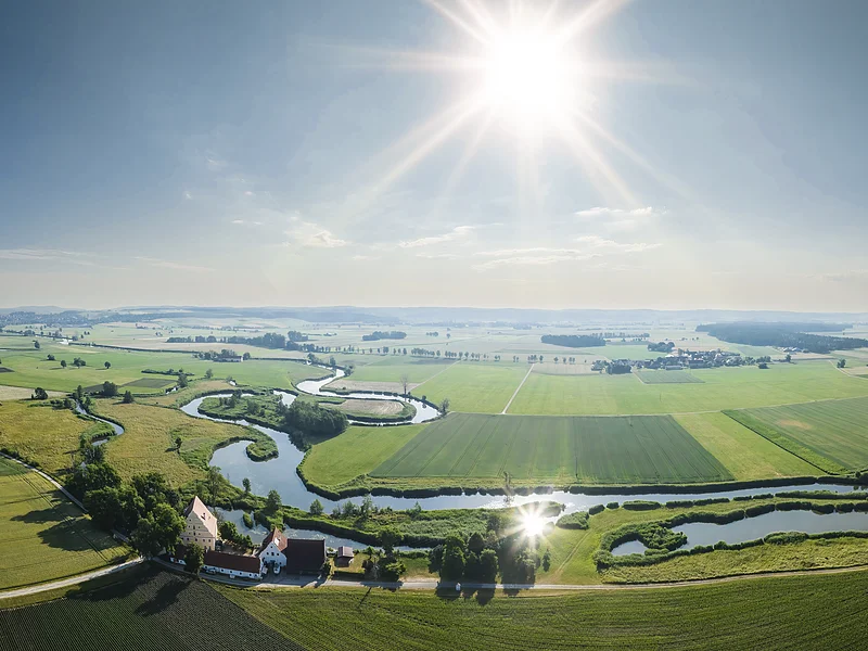 Wörnitzschleifen bei Munningen Landschaft mit einem sich schlängelndem Fluss, Feldern und Gebäuden, unter einem klaren Himmel mit strahlender Sonne.