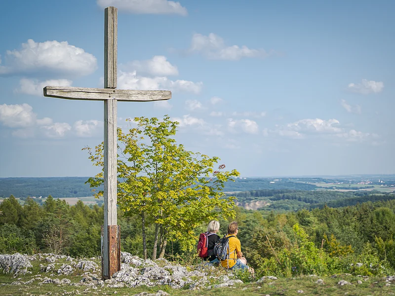Holzkreuz auf dem Harburger Bockberg, zwei Personen mit Rucksäcken sitzen daneben, Blick auf die bewaldete Landschaft.