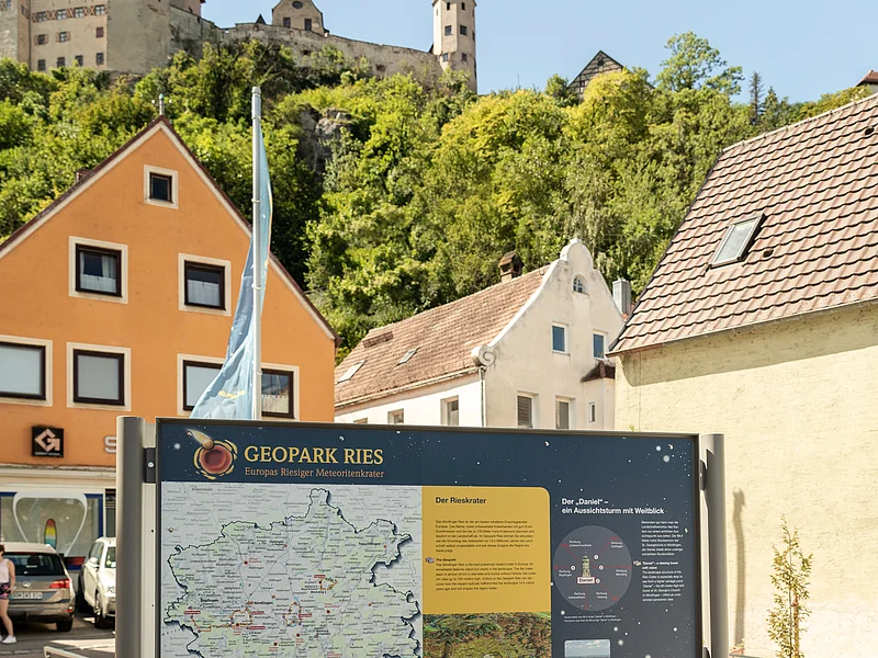 Unterhalb einer Burg steht zwischen Häusern eine Informationstafel über den Geopark Ries.