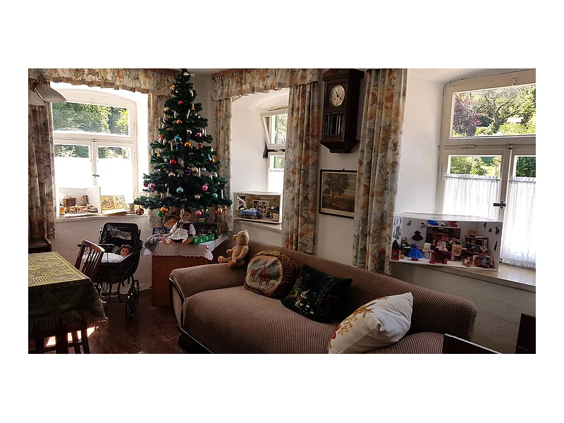 Wohnzimmer mit Weihnachtsbaum, Sofa, Puppen, Puppenwagen und Fenstern mit Gardinen und Blick ins Grüne