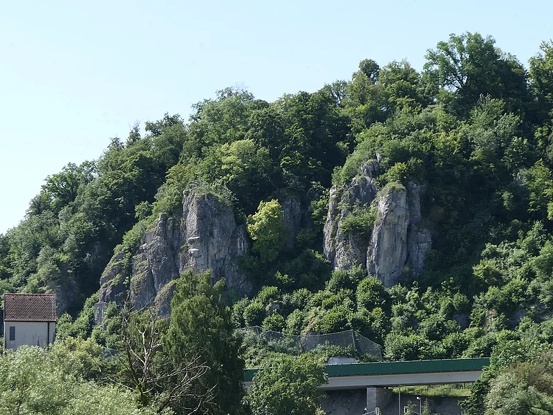 An einem mit Bäumen bewachsenen Hang befinden sich zwei rundliche Felsformationen. Im Vordergrund sind zwei Brücken und ein Fluss zu sehen.